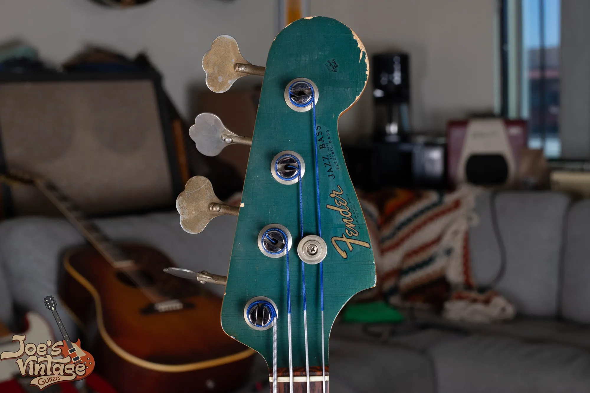 Matching painted headstock of a 1966 Fender Jazz Bass in Lake Placid Blue that has aged toward teal green, showing the gold Fender Jazz Bass transition logo, heavy lacquer checking, and edge wear through to the wood.